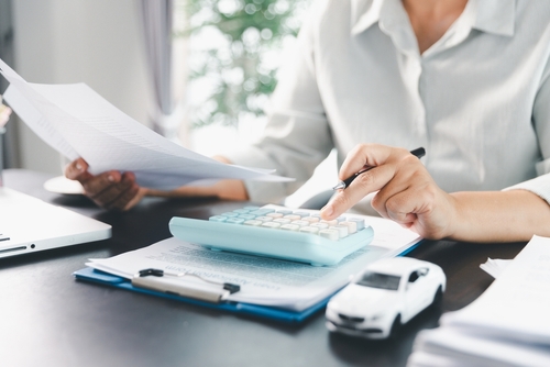 Person using a calculator while reviewing paperwork beside a miniature white car, illustrating the process of checking car finance costs and preparing a complaint.